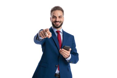 Mavi takım elbiseli mutlu işadamı kamerayı işaret ediyor ve elinde bir akıllı telefon tutarken gülümsüyor ve beyaz arka planda ileriye bakıyor.