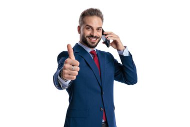 Approving businessman talking on smartphone and showing thumbs up while looking forward on white background