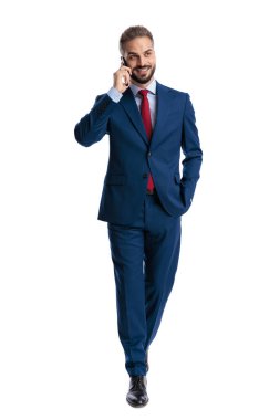 Cheerful businessman in a blue suit walking and talking on a mobile phone with hand in pocket on white background