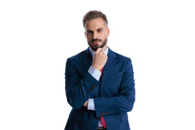 Portrait of a skeptical man wearing a formal blue suit touching chin and thinking while looking forward isolated on white background