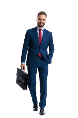 Confident young businessman in blue suit walking forward with hand in pocket and holding briefcase on white background