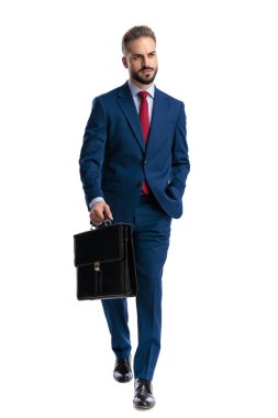 Determined young businessman in blue suit walking forward with briefcase and looking to side on white background