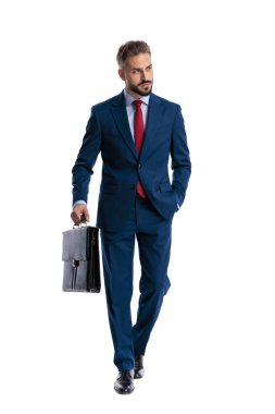 Serious businessman in blue suit walking forward with hand in pocket and holding briefcase while looking away on white background
