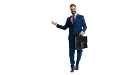 Mavi takım elbiseli mutlu genç adam beyaz arka planda yürürken bavul tutuyor ve yan yana sunum yapıyor.