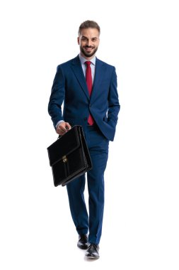 Confident businessman in blue suit holding briefcase and walking forward with hand in pocket while smiling and looking forward on white background