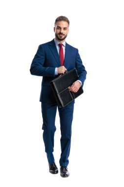 Confident businessman wearing blue suit walking forward holding leather briefcase with both hands and looking forward isolated on white background