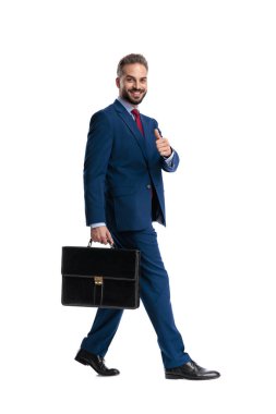 Side view of confident businessman holding briefcase walking showing thumbs up sign and looking forward isolated on white background