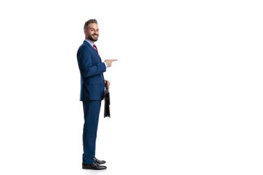 Mavi takım elbiseli, gülümseyen iş adamının fotokopi alanını göstermesi ve beyaz arka planda izole edilmiş evrak çantasını tutması.