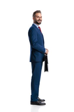 Side view of cheerful businessman wearing blue suit holding briefcase and standing isolated on white background
