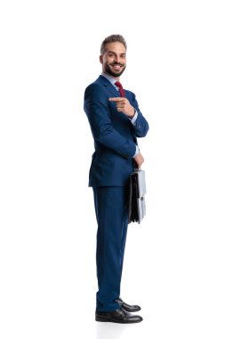 Side view of happy businessman wearing blue suit holding briefcase and pointing finger over shoulder at copy space while standing isolated on white background