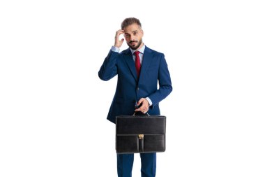 Kafası karışmış, mavi takım elbiseli, elinde siyah evrak çantası olan, başını kaşıyan beyaz arka planda ileriye bakan bir adam.