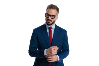 Zarif iş adamı gözlük takıyor ve mavi takım elbise giyiyor. Kollarını düzeltiyor ve beyaz arka planda yer almaya çalışıyor.