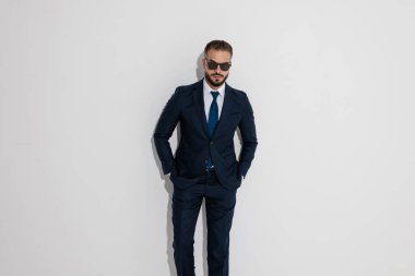 Serious man in a dark suit and sunglasses standing with hands in pockets looking forward on white background