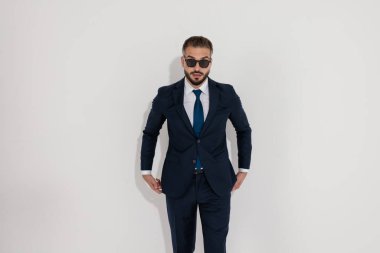 Mysterious businessman in a suit and sunglasses standing with hands near pockets while looking forward on white background