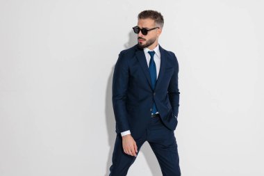 Confident man wearing a formal blue suit and sunglasses standing with hand in pocket while looking to the side on white background