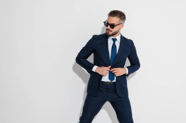 Elegant man wearing suit and sunglasses adjusting jacket and looking to side on white background