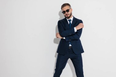 Cool man wearing blue suit and sunglasses posing with crossed arms and looking forward on white background