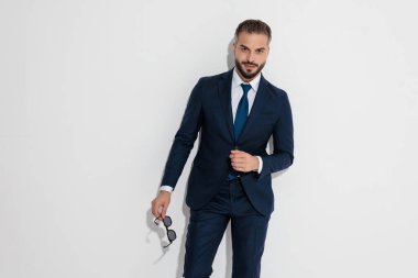 Confident man wearing blue suit and tie holding sunglasses while looking forward over white background