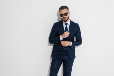 Elegant man wearing blue suit and sunglasses adjusting sleeve while looking forward over white background