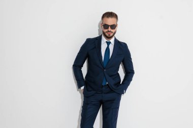 Stylish man wearing suit and sunglasses standing with hands in pockets while looking forward over white background