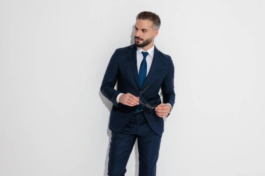 Elegant businessman wearing blue suit holding sunglasses while looking to side against white background