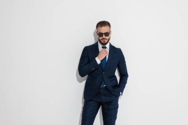 Confident businessman wearing sunglasses and adjusting tie while standing while looking forward with hand in pocket on white studio background