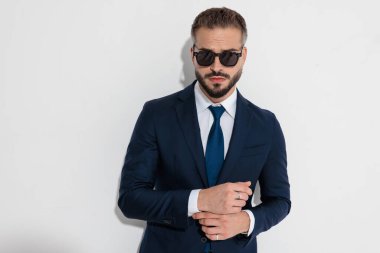 Tough man in navy suit wearing sunglasses and adjusting sleeves while looking forward against white background