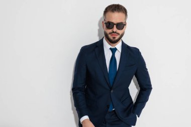 Confident man wearing navy suit and sunglasses with hand in pocket while looking forward against white background