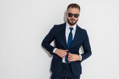 Elegant man wearing navy suit and sunglasses adjusting jacket while looking forward against white background