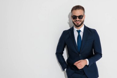 Cool businessman wearing blue suit and sunglasses smiling while standing with hand in pocket against white background