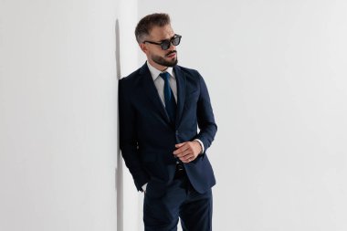 Tough businessman wearing blue suit and sunglasses looking to side while leaning on wall against white background