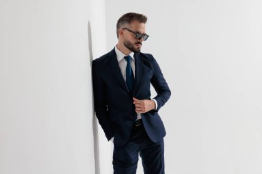 Cool man wearing blue suit and sunglasses adjusting jacket while leaning on wall and looking down against white background