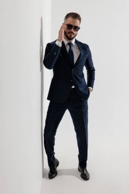 Serious man wearing blue suit and sunglasses adjusting glasses while leaning against wall with hand in pocket on white background