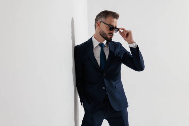 Mysterious man wearing blue suit adjusting sunglasses while leaning against wall on white background