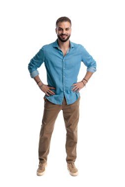 Kendine güvenen, kot gömlek giyen ve elleri belinde gülümseyen genç bir adam beyaz arka planda tek başına ayakta ve ileriye bakıyor.