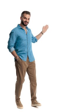 Kot gömlekli, gülümseyen, cebinde el ele duran ve beyaz arka planda ileriye bakarak el kol hareketi yapan bir adam.