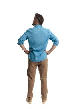 Kot gömlek giyen ve kahverengi pantolon giyen, elleri kalçasında duran ve beyaz arka planda izole bir şekilde yukarı bakan kendine güvenen bir adam.