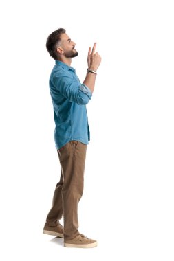 Heyecanlı sıradan bir adamın yukarı bakıp yukarı doğru işaret ederken beyaz arka planda mavi gömlek ve kahverengi pantolon giymesi.