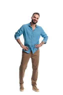 Kendine güvenen, gülümseyen ve elleri kalçasında duran, beyaz arka planda yalnız bir şekilde ileriye bakan bir adam.