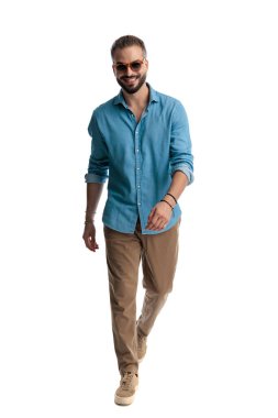 Kot gömlek ve güneş gözlüğü takan şık bir adam ileri doğru yürüyor ve beyaz arka plana bakarken gülümsüyor.