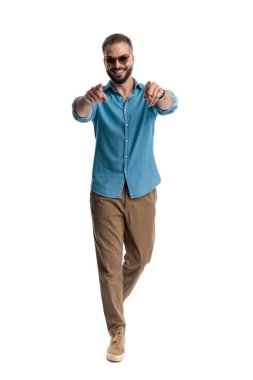 Güneş gözlüklü şık sakallı adam yürüyor ve gülümsüyor ve iki elini de beyaz arka planda izole edilmiş kameraya doğrultuyor.