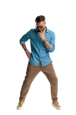 Düşünceli bir adam güneş gözlüğü ve kot gömlek takıyor dudağa dokunuyor ve beyaz arka planda poz veriyor.