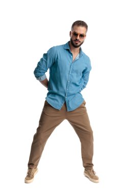 Havalı, güneş gözlüğü takan, elleri arkada duran ve beyaz arka planda ileri bakan bir adam.