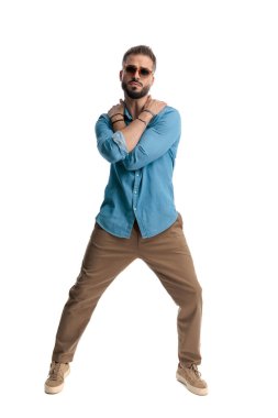 Kot gömlek ve güneş gözlüğü takan ciddi bir adam. Bacakları açık, kolları göğsünün üstünde beyaz bir arkaplan ararken kollarını kavuşturuyor.