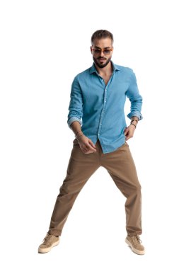 Sakallı ve burun halkalı, rahat kıyafetler ve güneş gözlüğü takan, geniş duruşlu ve beyaz arka planda ileriye bakan kendinden emin bir adam.