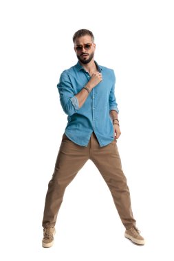 Güneş gözlüğü ve kot gömlek takan havalı bir adam hazırlanırken yakasını düzeltiyor ve beyaz arka planda ileriye bakıyor.