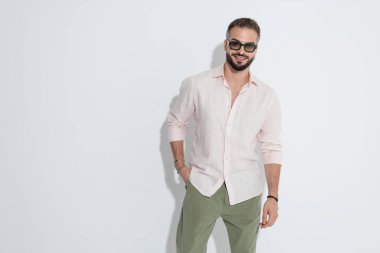 Güneş gözlüğü ve pembe gömlek takan şık sakallı adam cebinde duruyor ve beyaz arka plana bakıyor.