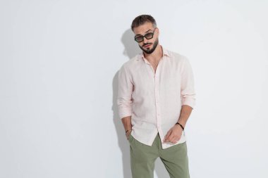 Güneş gözlüğü ve pembe gömlek takan yakışıklı sakallı adam el ele duruyor ve beyaz arka plana bakıyor.