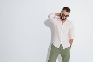 Burun halkalı, gözlüklü ve pembe tişörtlü mutlu sakallı bir adam. El ele tutuşmuş, boynuna dokunuyor ve beyaz arka planda gülümseyerek aşağı bakıyor.