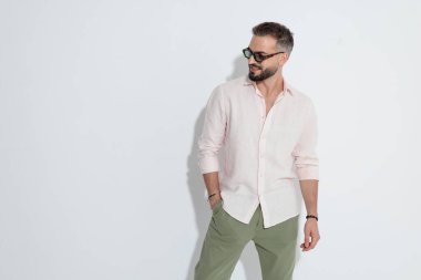 Burun halkası takan, pembe gömlekli, güneş gözlüklü, el ele duran, beyaz arka planda mutlu bir gülümseme arayan modaya uygun sakallı bir adam.